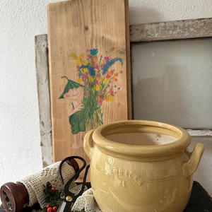 Puede incluir: Una olla de cerámica amarilla con dos asas, una tabla de madera con una ilustración floral y unas tijeras negras. La olla tiene un esmalte craquelado y la tabla presenta a un niño sosteniendo flores. También hay una bobina de madera y encaje.