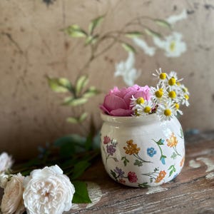 Könnte beinhalten: Eine kleine, weiße Keramikvase mit bunten Blumenmustern, die eine rosa Rose und kleine weiße und gelbe Blumen enthält. Die Vase steht auf einer Holzoberfläche, mit zusätzlichen Rosen in der Nähe.