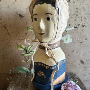 Könnte beinhalten: Eine dekorative Pappmaché-Büste einer Person mit bemalten Gesichtszügen, die eine cremefarbene Spitzenhaube trägt, welche mit Bändern unter dem Kinn gebunden ist. Die Büste hat ein dunkelblaues Oberteil mit goldfarbenen Akzenten auf einem runden blauen Sockel.