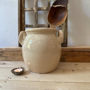 Könnte beinhalten: Eine cremefarbene Keramik-Vase mit zwei Henkeln. Die Vase hat ein dekoratives Design auf der Vorderseite. Die Vase steht auf einer Holzoberfläche mit einem Holzlöffel und einer kleinen Kerze.