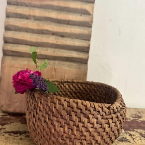 May include: A small, round, brown woven basket with a thick, textured rim. A single bright pink flower and a small sprig of purple lavender with green leaves are tucked into the side of the basket, resting against the woven material.