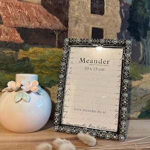 May include: A silver picture frame with a pearl-like trim and a white mount. The frame is sitting on a wooden surface with a vase of flowers and dried grasses in the background. The frame has the text "Meander elegant home decorations 10 x 15 cm www.meander-bv.nl" printed on the mount.