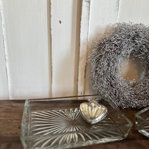 Puede incluir: Un plato cuadrado de vidrio transparente con un patrón decorativo de rayos de sol en el fondo. Un adorno en forma de corazón plateado está sentado dentro del plato.