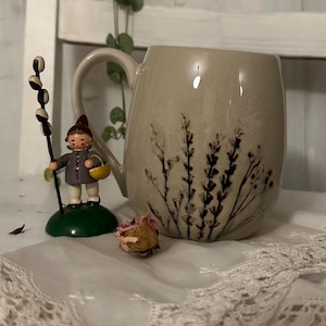 May include: A beige ceramic mug with a floral design. The mug has a rounded shape and a handle. The mug is sitting on a white lace doily.