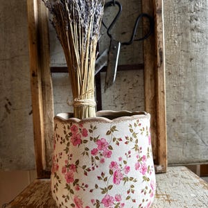 Peut inclure: Un pot en céramique avec un bord festonné et une finition émaillée blanche craquelée, orné d'un motif répétitif de petites fleurs roses et de feuilles vertes. Un bouquet de tiges de lavande séchée est placé à l'intérieur.
