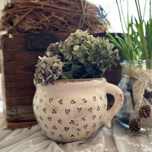 Op de afbeelding: Een witte keramische pot met een handvat, versierd met een patroon van kleine zwarte hartjes. De pot is gevuld met gedroogde bloemen en staat op een wit kantkleed.