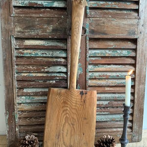 Könnte beinhalten: Ein großes, rustikales Holzbrett zum Schneiden mit einem langen Griff. Das Brett ist aus hellem braunem Holz mit natürlicher Maserung. Es lehnt an einem verwitterten Holzladen.