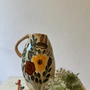 Pode incluir: Um jarro de cerâmica com base em tom creme, apresentando motivos florais pintados à mão em amarelo, castanho e verde. O design inclui um girassol amarelo proeminente e uma flor castanha escura, rodeados por folhagem verde e espigas de trigo.
