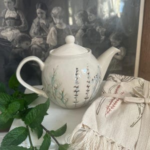 Peut inclure: Théière en céramique crème avec un couvercle, ornée d'un motif floral bleu et vert. Un torchon assorti avec un motif floral similaire et des franges est à côté de la théière. Des brins de menthe fraîche sont au premier plan.