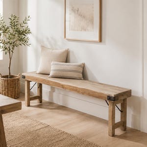 Op de afbeelding: Een lange, rechthoekige houten bank met een natuurlijke lichtbruine afwerking, voorzien van donkere metalen hoekbeugels op de zitting en vier stevige poten. Twee rechthoekige sierkussens van beige en gestreepte stof liggen op de bank.