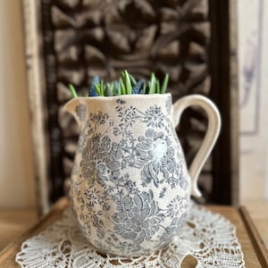 以下が含まれることがあります： 青い花柄の白い陶器のピッチャーに、青と緑の花の小さなブーケが入っています。ピッチャーは白いレースのドイリーの上に置かれています。