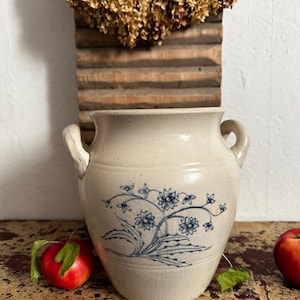 Può includere: Un vaso in ceramica beige con due manici laterali e un disegno floreale blu sulla parte anteriore. Il recipiente presenta una finitura liscia e smaltata con un bordo largo e arrotondato. Il motivo blu raffigura fiori e foglie stilizzati.