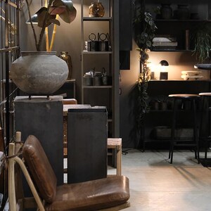 May include: A brown leather chair with a wooden frame sits in a room with a dark grey wall and a shelf unit with various items. A large grey pot with a plant sits on a grey pedestal.