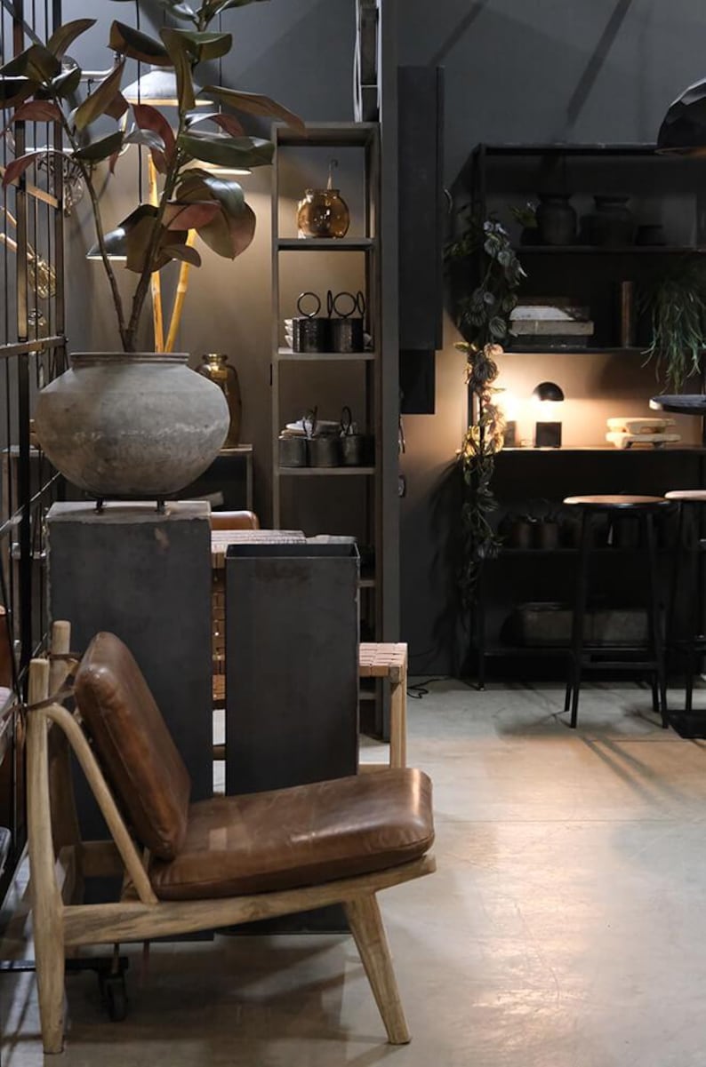 May include: A brown leather chair with a wooden frame sits in a room with a dark grey wall and a shelf unit with various items. A large grey pot with a plant sits on a grey pedestal.