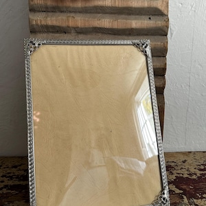 May include: A rectangular picture frame featuring a metallic silver border with an intricate embossed pattern and decorative corner accents. The frame contains a clear glass pane covering a plain, light beige, textured paper insert.