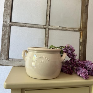 Może przedstawiać: Kremowy ceramiczny garnek z dwoma uchwytami i wytłoczonym tekstem "Drobacka 2L" na boku. Garnek stoi na jasnożółtym drewnianym stole z szufladą. Obok garnka stoi bukiet fioletowych kwiatów bzu.