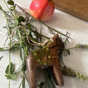 Pode incluir: Uma figura de cavalo Dala de madeira com uma sela verde e detalhes pretos repousa entre raminhos de ervas e uma maçã vermelha. O cavalo é castanho com um desenho pintado. A maçã é vermelha e amarela. O fundo é uma superfície de madeira branca.
