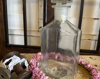 May include: A clear glass bottle with a square shape and a glass stopper. The bottle is sitting on a wooden surface with a white lace doily and a pink flower garland.