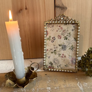 Könnte beinhalten: Eine brennende weiße Kerze in einem blütenförmigen Messingkerzenhalter neben einem goldgerahmten Bild mit Blumendruck. Der Rahmen ist mit kleinen Perlen verziert. Rechts ist eine getrocknete grüne Pflanze.