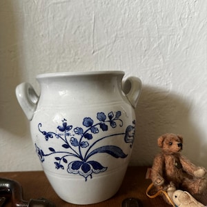 Könnte beinhalten: Ein weißer Keramiktopf mit zwei seitlichen Henkeln, verziert mit einem blauen Blumenmuster aus Blättern und Blüten. Der Topf hat einen bauchigen Körper und einen ausgestellten Rand. Daneben sitzt ein kleiner brauner Plüschteddy auf einem Spielzeugwagen.