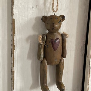 Puede incluir: Adorno de oso de peluche de madera marrón con un corazón pintado en el pecho. El oso tiene una cuerda para colgar. El fondo es una superficie blanca y texturizada. El oso es de madera.