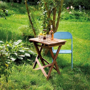 Op de afbeelding: Een houten klaptafel met een blauwe klapstoel op een grasveld. Op de tafel staan een fles wijn en glazen.