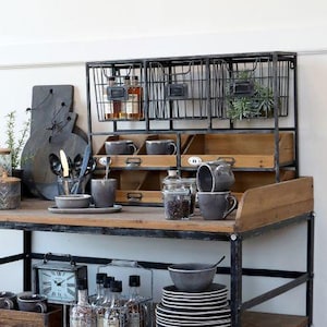 Op de afbeelding: Een rustieke, industriële keukenopslagunit met draadmandjes, houten lades en een metalen frame. De unit toont diverse keukenspullen, waaronder mokken, borden en potten, wat een functionele en decoratieve presentatie creëert.
