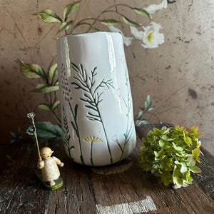 Könnte beinhalten: Eine weiße Keramikvase mit geprägten und grün bemalten botanischen Illustrationen von Wildblumen und Gräsern. Die Vase hat eine leicht konische, zylindrische Form mit einer glatten, glänzenden Oberfläche und einem dunkelgrünen Rand oben.