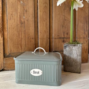 Puede incluir: Una panera verde salvia con un asa blanca y una etiqueta ovalada blanca que dice "Brot". La caja tiene un diseño acanalado y una forma rectangular. Una flor blanca en una maceta de piedra está a la derecha.