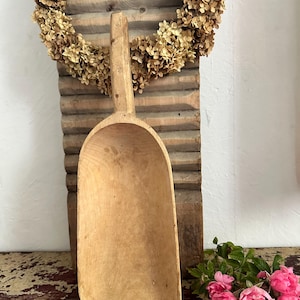 Puede incluir: Una pala de madera de color marrón claro con un mango largo y un cuenco profundo y redondeado. La pala presenta una textura de veta de madera natural y suave, con un diseño sencillo y funcional. Está tallada en una sola pieza de madera.