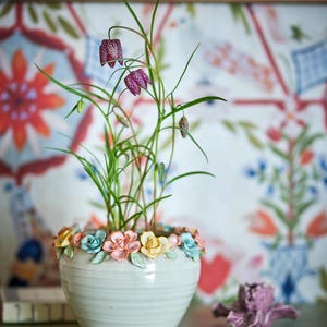 Op de afbeelding: Een witte keramische bloempot met een bloemenontwerp rond de rand, met een plant met groene stelen en paarse, geblokte bloemen. De pot staat op een groen oppervlak, met een decoratieve achtergrond.