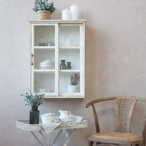 Puede incluir: Un mueble de pared blanco, de aspecto envejecido, con puertas de cristal, que muestra tazas, tarros y utensilios blancos. Una pequeña mesa sostiene una bandeja con tazas y un jarrón. Una silla de madera con respaldo tejido está a la derecha. Velas decorativas y una planta encima.