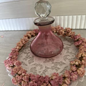 Könnte beinhalten: Eine rosa Glasflasche mit einem facettierten Glasstopfen steht auf einem weißen Spitzenuntersetzer, umgeben von einem Kranz aus getrockneten rosa Blüten.