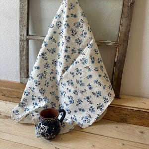 May include: A white fabric with a blue floral pattern hangs from a hook on a rustic wooden window frame. A blue and white ceramic pitcher sits on a wooden floor.
