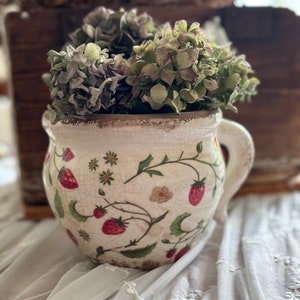 Könnte beinhalten: Ein weißer Keramik-Krug mit Blumenmuster und Henkel, gefüllt mit getrockneten Hortensien. Der Krug steht auf einer weißen Spitzentischdecke.