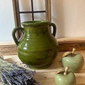 Pode incluir: Um vaso de cerâmica verde com duas alças, ao lado de duas figuras de maçã de cerâmica verde com caules e folhas de madeira. Um feixe de lavanda seca e uma moldura de janela de madeira também estão presentes.
