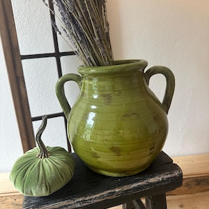 May include: A green ceramic vase with two handles, filled with dried lavender. A small, green velvet pumpkin sits on a distressed black wooden stool. The vase has a glossy finish and a rustic appearance.