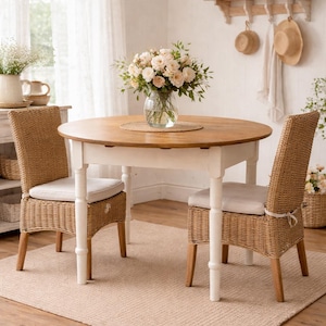 Op de afbeelding: Een ronde houten eettafel met een witte basis en twee rieten stoelen met crèmekleurige kussens. Een vaas met bloemen staat op de tafel. De kamer heeft een neutraal kleurenpalet met een vloerkleed en houten vloer.