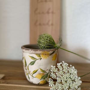 Op de afbeelding: Een witte keramische pot met een citroen- en bladontwerp. De pot staat op een houten oppervlak met een witte bloem op de voorgrond.