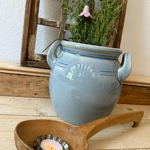 May include: A blue ceramic pot with the text "ROBACK SL" on the side, filled with dried grasses and a pink ceramic flower. A wooden spoon with a metal tea light holder in the bowl.