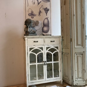 Könnte beinhalten: Weißer, abgenutzter Schrank mit Glastüren und zwei Schubladen. Der Schrank hat eine abgenutzte weiße Oberfläche und ein dekoratives Bogenmuster auf den Glastüren. Der Schrank steht auf einem Holzboden.