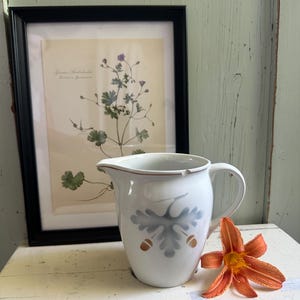 以下が含まれることがあります： グレーのオークの葉のモチーフと2つの小さな茶色のどんぐりが描かれた白い陶器製のクリーマーピッチャー。ピッチャーには湾曲したハンドル、縁に小さな欠けのある注ぎ口、そして容器の上端を縁取る細い茶色のラインがあしらわれています。
