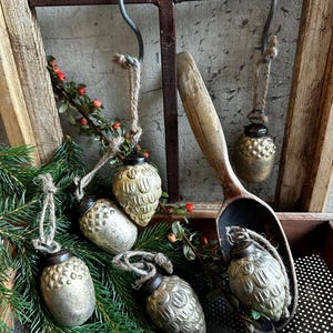 Op de afbeelding: Vijf vintage-stijl kwikglas ornamenten in de vorm van dennenappels en eikels, met metallic zilveren en gouden afwerkingen, donkere metalen dopjes en lussen van jutekoord. Eén ornament ligt in een verweerde houten schep bij dennentakken.