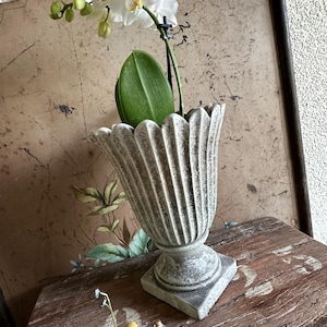Könnte beinhalten: Eine weiße Orchidee in einem grauen, geriffelten urnenförmigen Pflanzgefäß. Der Pflanztopf steht auf einer Holzoberfläche mit einer kleinen Figur eines Kindes, das eine Blume hält. Der Hintergrund zeigt eine Wanddekoration im Vintage-Stil.