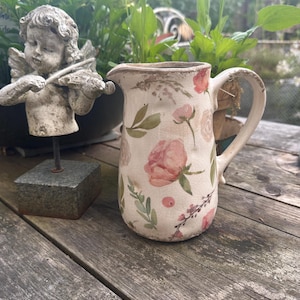 Può includere: Una brocca in ceramica con un motivo floreale di rose rosa e foglie verdi. La brocca ha un manico e uno sfondo bianco sporco leggermente invecchiato. La brocca è appoggiata su una superficie di legno usurata.