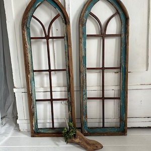 Könnte beinhalten: Zwei rustikale, bogenförmige Fensterrahmen aus Holz mit abgenutzter türkis- und brauner Farbe. Die Rahmen haben ein Metallgitterdesign. Ein Holzschuhleisten mit Grünzeug befindet sich davor.