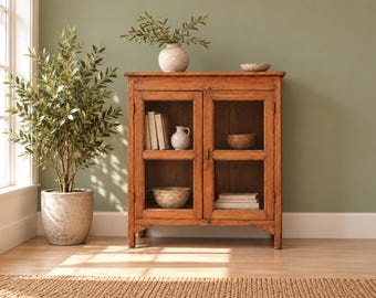 Puede incluir: Un armario de madera con dos puertas de cristal, con un acabado marrón natural. En el interior, los estantes contienen libros, una pequeña jarra de cerámica y dos cuencos. Un olivo en maceta se encuentra a la izquierda.