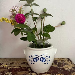 Könnte beinhalten: Ein weißer Keramiktopf mit zwei seitlichen Griffen und einem blau bemalten Blumenmotiv auf der Vorderseite, gefüllt mit einem Strauß aus grünen Stielen, rosa Blüten, gelben Blumen und drei grünen Mohnkapseln in einem Glaseinsatz.