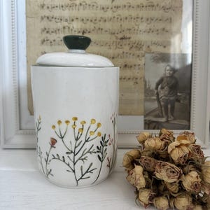 Puede incluir: Jarrón de cerámica blanco con tapa verde y diseño floral. El jarrón tiene un diseño floral con flores y hojas amarillas y verdes. El jarrón está sobre una superficie blanca.