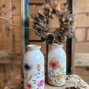Könnte beinhalten: Zwei weiße Keramikvasen mit floralen Designs. Eine Vase zeigt blaue, rosa und orangefarbene Blumen, die andere rote und gelbe Blüten. Die Vasen haben eine verwitterte, geknackte Oberfläche und stehen auf einer Holzoberfläche.